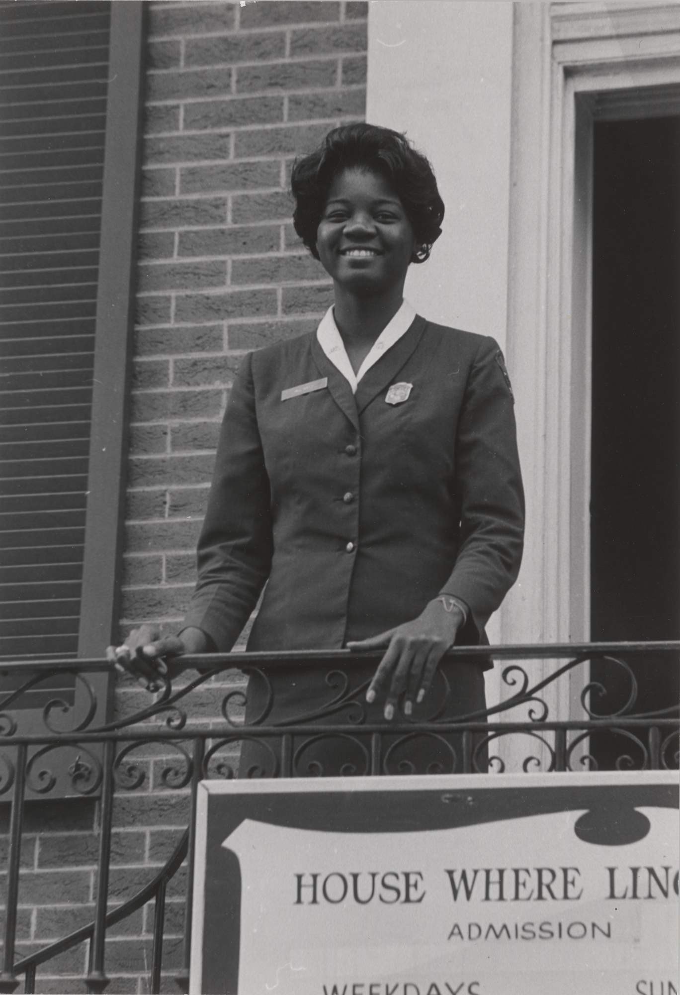 Miss Rolle stands smiling at a railing on top of steps. She wears the NPS women’s uniform skirt, blouse, and jacket with shield-shaped badge.